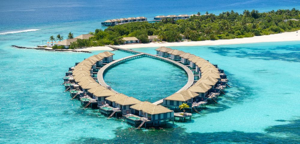 Aerial view of luxury overwater villas in a heart shape, surrounded by turquoise waters and lush tropical vegetation.