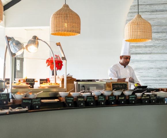 Chef in white standing in front of a array of buffet breakfast foods with wicker lights above