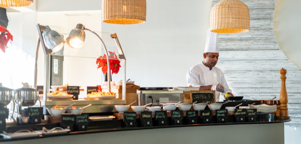 Chef in white standing in front of a array of buffet breakfast foods with wicker lights above