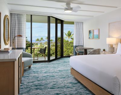 A bright hotel room featuring a queen bed, modern furnishings, and a balcony with ocean views and tropical foliage.