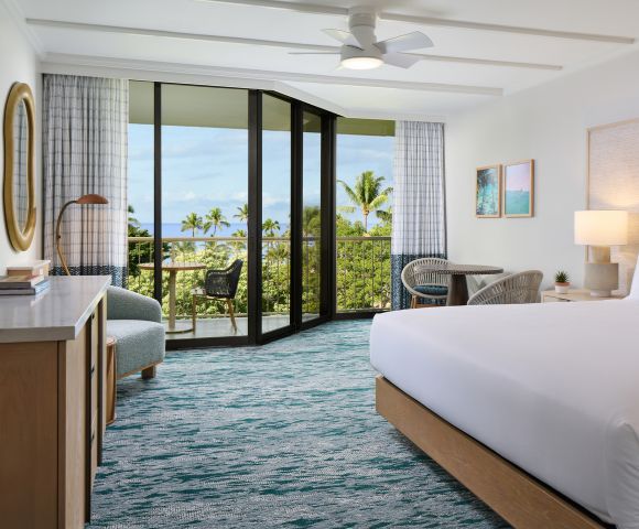 A bright hotel room featuring a queen bed, modern furnishings, and a balcony with ocean views and tropical foliage.