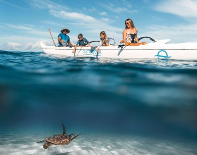 A group of four people is exploring the ocean from an outrigger canoe on a sunny day, surrounded by clear water and blue skies.