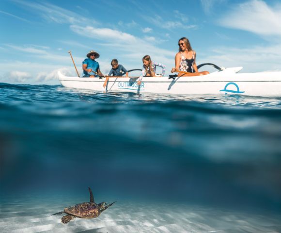 A group of four people is exploring the ocean from an outrigger canoe on a sunny day, surrounded by clear water and blue skies.
