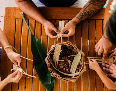 Aerial view of hands weaving palm leaves into a basket on a wooden table, with a green leaf and playful gestures from children.
