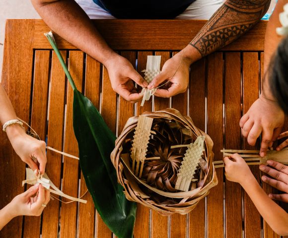 Aerial view of hands weaving palm leaves into a basket on a wooden table, with a green leaf and playful gestures from children.