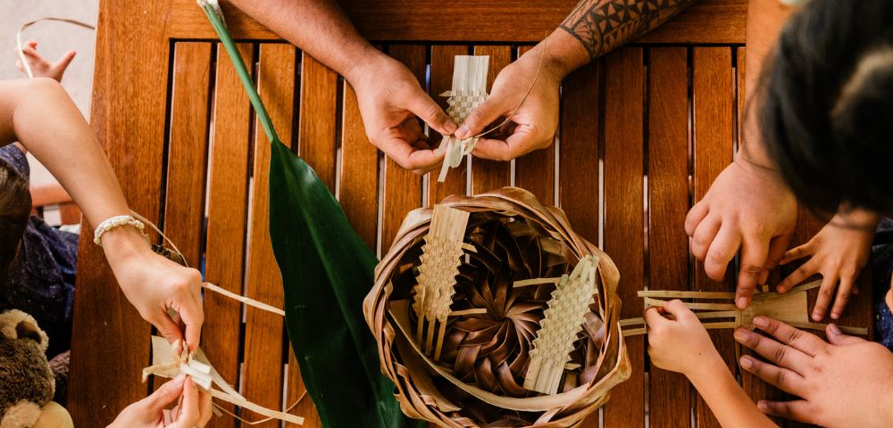 Aerial view of hands weaving palm leaves into a basket on a wooden table, with a green leaf and playful gestures from children.