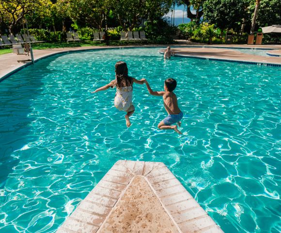 Two children joyfully leap into a bright blue swimming pool from a diving board. Lush greenery surrounds the pool area.
