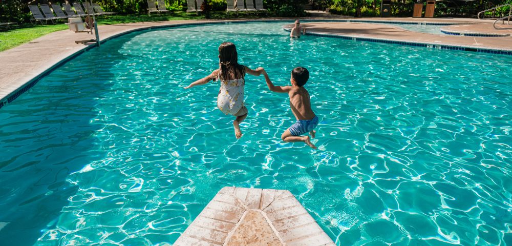 Two children joyfully leap into a bright blue swimming pool from a diving board. Lush greenery surrounds the pool area.
