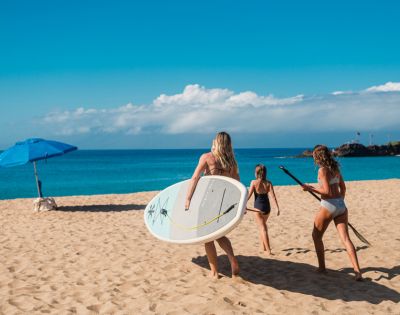 Three girls walk along a sandy beach, carrying a surfboard and paddle, under a sunny sky with an umbrella nearby and ocean waves ahead.
