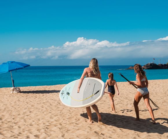 Three girls walk along a sandy beach, carrying a surfboard and paddle, under a sunny sky with an umbrella nearby and ocean waves ahead.