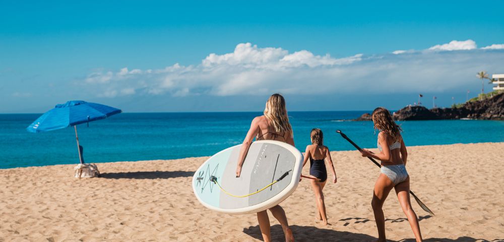 Three girls walk along a sandy beach, carrying a surfboard and paddle, under a sunny sky with an umbrella nearby and ocean waves ahead.