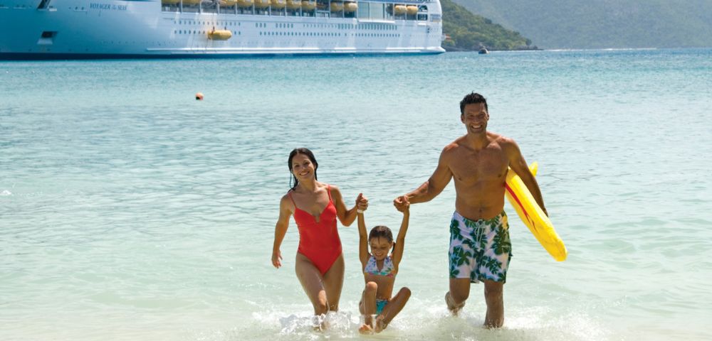 A family wades in clear water, with a large cruise ship docked in the background amidst green hills under a blue sky.