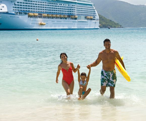 A family wades in clear water, with a large cruise ship docked in the background amidst green hills under a blue sky.