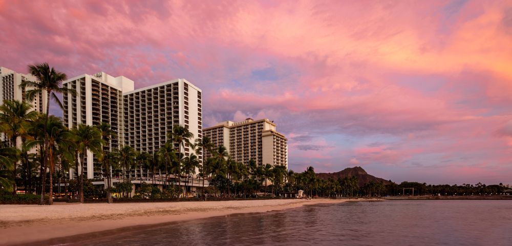 Sunset beach view featuring tropical palm trees, hotels, and a vibrant pink sky over calm waters. Diamond Head mountain in the distance.