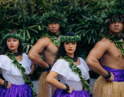 A group of dancers in traditional Hawaiian attire, featuring purple skirts and leafy garlands, standing confidently amidst lush greenery.