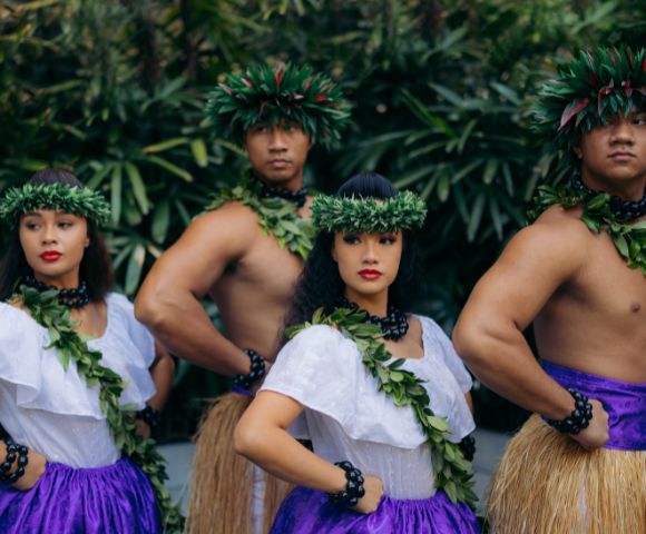 A group of dancers in traditional Hawaiian attire, featuring purple skirts and leafy garlands, standing confidently amidst lush greenery.