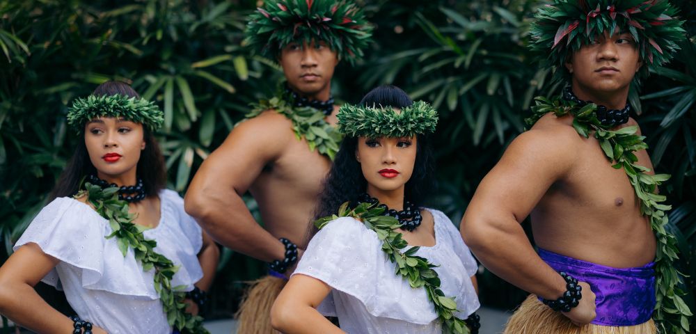 A group of dancers in traditional Hawaiian attire, featuring purple skirts and leafy garlands, standing confidently amidst lush greenery.