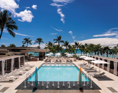 A sunny pool area at a resort, surrounded by palm trees, lounge chairs, and umbrellas, with a view of the beach and blue sky.