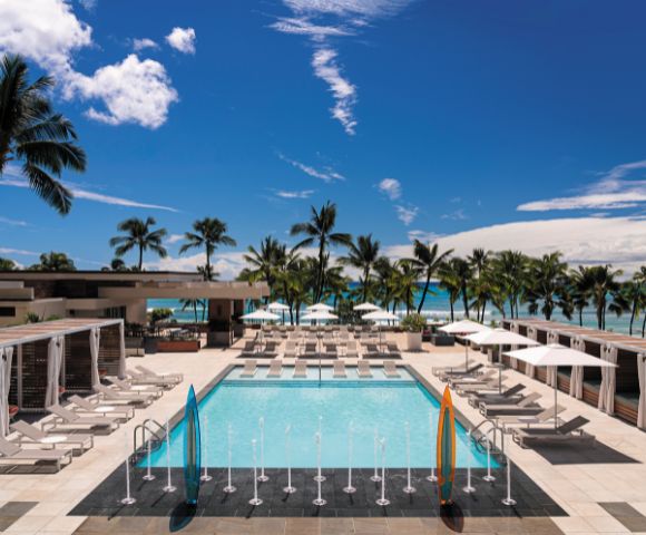 A sunny pool area at a resort, surrounded by palm trees, lounge chairs, and umbrellas, with a view of the beach and blue sky.