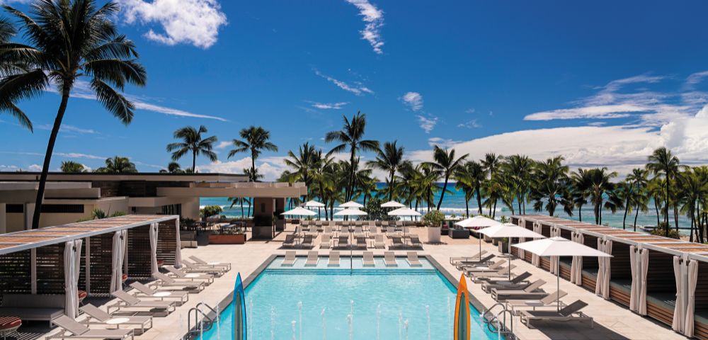 A sunny pool area at a resort, surrounded by palm trees, lounge chairs, and umbrellas, with a view of the beach and blue sky.
