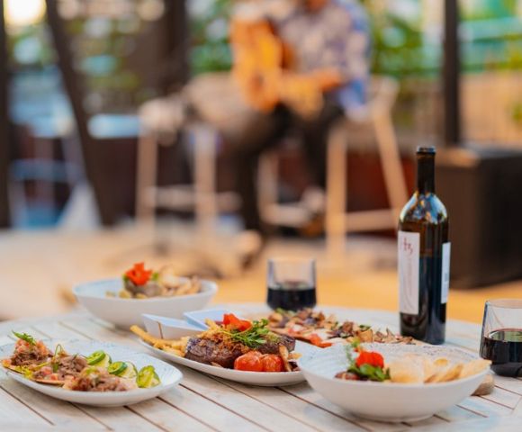 Dining table with a variety of dishes, red wine, and glasses, with a musician playing guitar in the background.