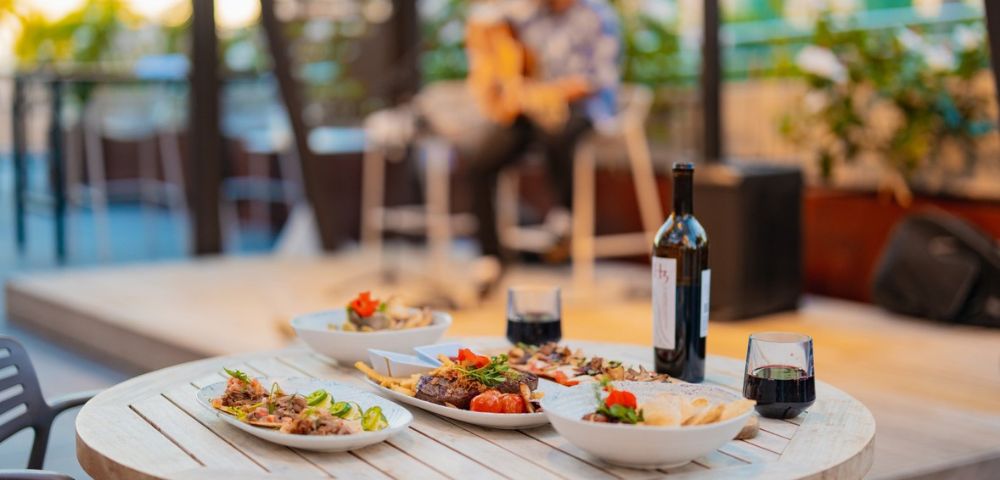 Dining table with a variety of dishes, red wine, and glasses, with a musician playing guitar in the background.