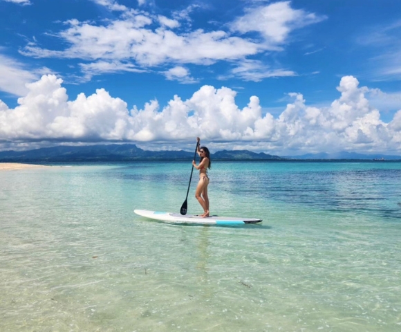 A woman paddles on a stand-up paddleboard in clear turquoise waters under a blue sky with fluffy clouds.