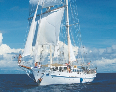A majestic white sailboat, the Spirit of the Pacific, glides through turquoise waters under a bright blue sky with fluffy clouds.