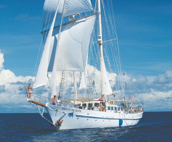 A majestic white sailboat, the Spirit of the Pacific, glides through turquoise waters under a bright blue sky with fluffy clouds.
