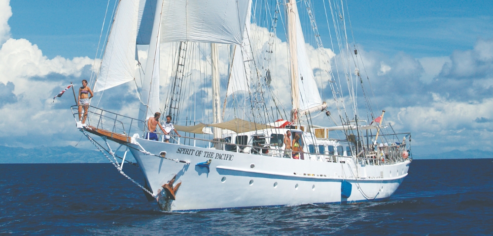 A majestic white sailboat, the Spirit of the Pacific, glides through turquoise waters under a bright blue sky with fluffy clouds.