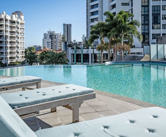 A serene rooftop pool surrounded by modern buildings and palm trees, with sun loungers in the foreground and clear blue skies above.