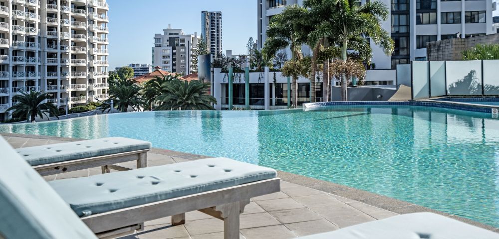 A serene rooftop pool surrounded by modern buildings and palm trees, with sun loungers in the foreground and clear blue skies above.