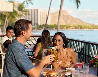 A couple enjoys drinks at a beachfront restaurant with palm trees and Diamond Head mountain in the background.