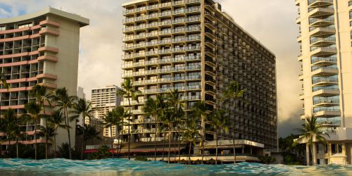 A view of high-rise hotels with palm trees in front, set against a cloudy sky and ocean waves in the foreground.