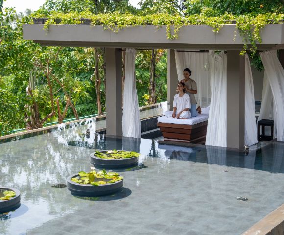 A tranquil outdoor spa setting features a massage table under a canopy, surrounded by lush greenery and calm water with lily pads.