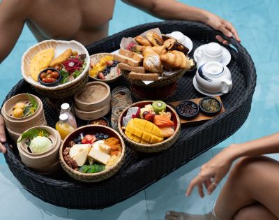 A stylish floating breakfast tray filled with fruits, pastries, cheese, and dim sum, enjoyed by a person in a pool.