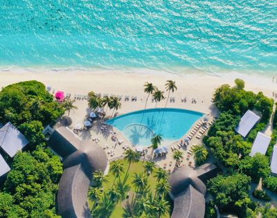 Aerial view of a tropical resort featuring a pool, sandy beach, turquoise ocean, and palm trees under bright sunlight.