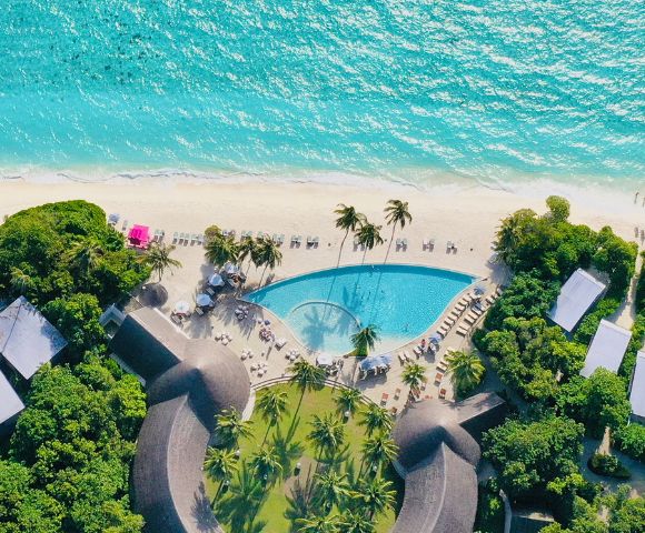 Aerial view of a tropical resort featuring a pool, sandy beach, turquoise ocean, and palm trees under bright sunlight.