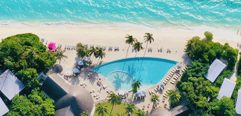Aerial view of a tropical resort featuring a pool, sandy beach, turquoise ocean, and palm trees under bright sunlight.