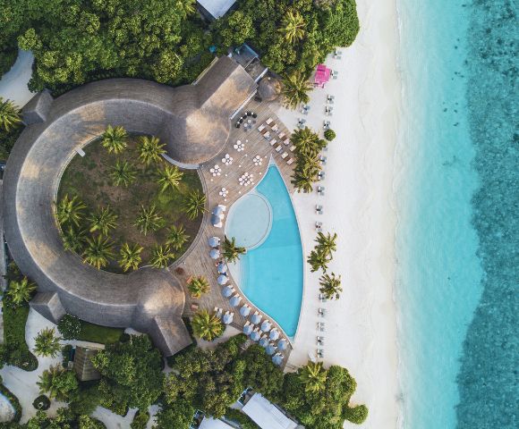 Aerial view of a tropical resort featuring a curved pool surrounded by palm trees, beach chairs, and pristine turquoise waters.
