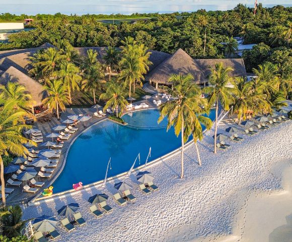 Aerial view of a tropical resort with a swimming pool, palm trees, and a sandy beach, surrounded by lush greenery and colorful cabanas.
