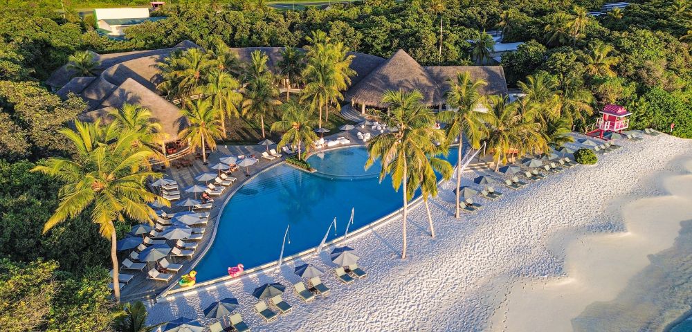 Aerial view of a tropical resort with a swimming pool, palm trees, and a sandy beach, surrounded by lush greenery and colorful cabanas.