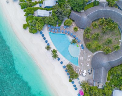 Aerial view of a tropical resort featuring a curved pool, sandy beach, palm trees, and lush greenery by clear turquoise waters.