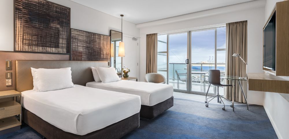 Bright hotel room with two twin beds, modern decor, a desk, and large windows offering a view of the ocean and outdoor seating.