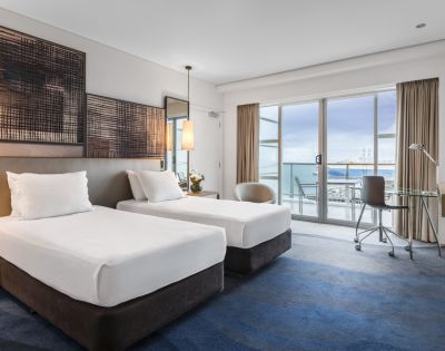 Bright hotel room with two twin beds, modern decor, a desk, and large windows offering a view of the ocean and outdoor seating.