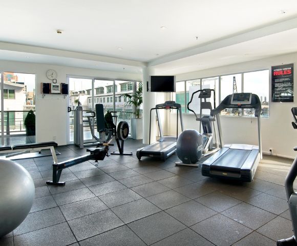 Bright, modern gym with cardio equipment, weights, and a large window view, featuring a clock and fitness center rules on the wall.