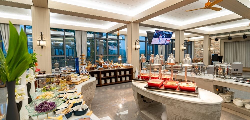 A spacious buffet spread with various dishes, glassware, and decorative plants, illuminated by warm lighting in a modern dining area.