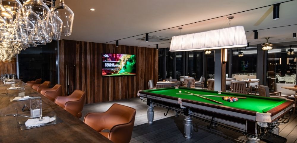 A stylish bar featuring a green pool table, modern bar seating, and an illuminated light fixture, with a TV displaying vibrant content.
