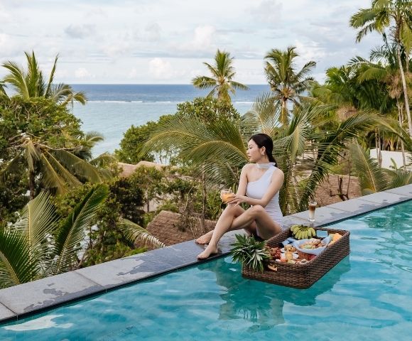 A woman in swimwear relaxes by a pool, holding a drink, with a basket of tropical fruits nearby and a scenic ocean view.