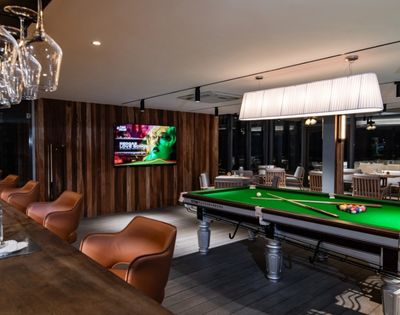 A stylish bar featuring a green pool table, modern bar seating, and an illuminated light fixture, with a TV displaying vibrant content.